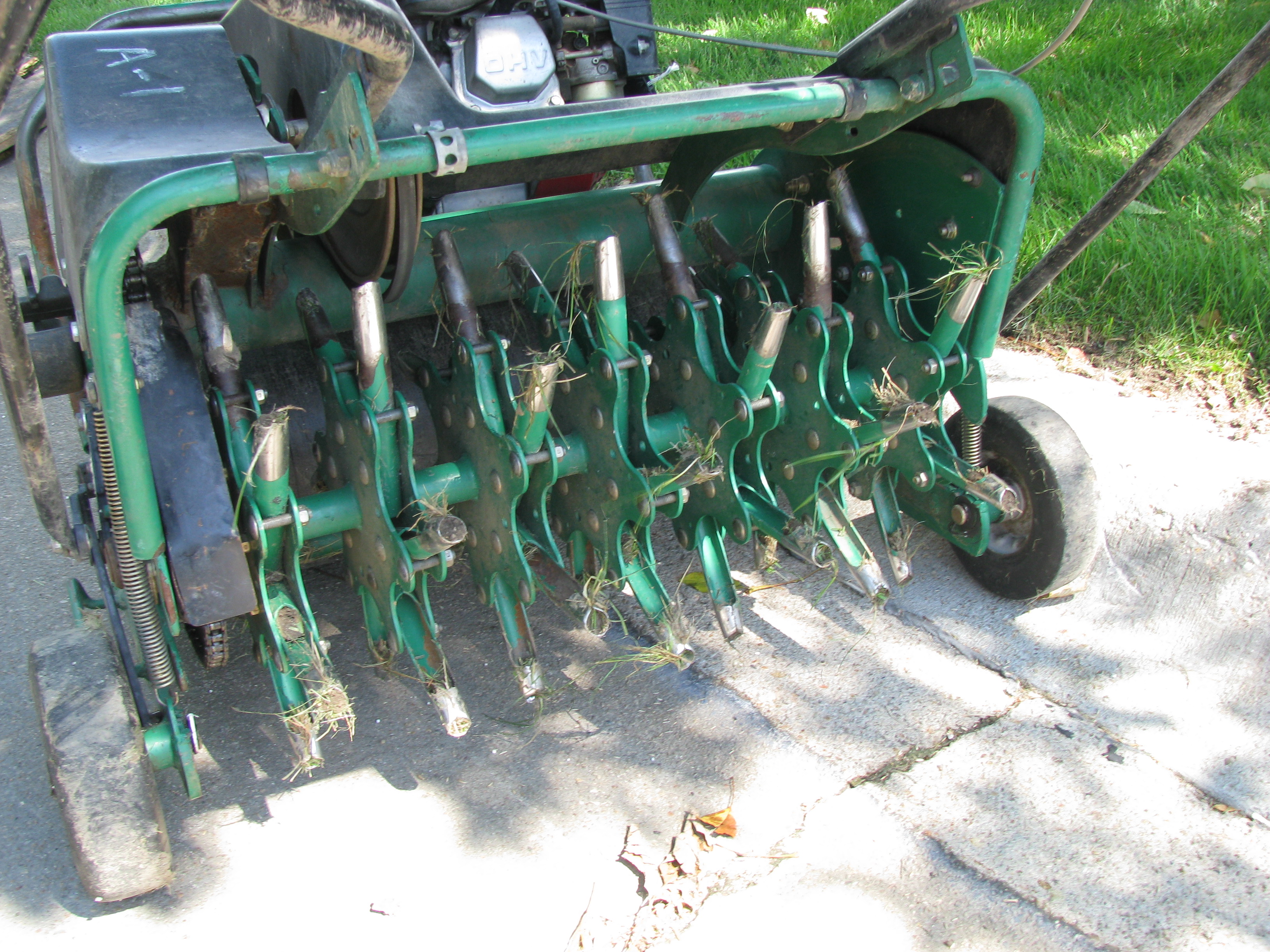 Lawn aerator machine with green hollow tines and small amounts of grass stuck on them.